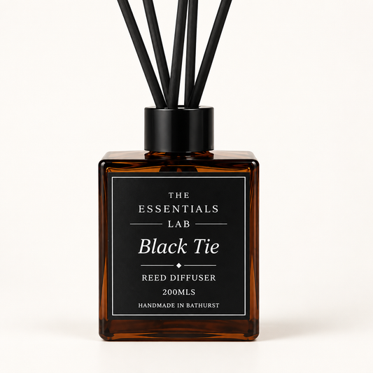 Amber glass reed diffuser bottle with a black cap and six black reeds, featuring a black label with white text that reads 'THE ESSENTIALS LAB Black Tie REED DIFFUSER 200MLS HANDMADE IN BATHURST' on a clean off-white background.