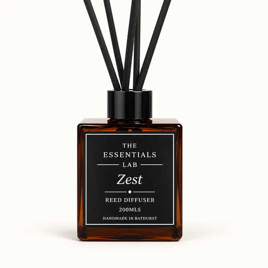 An amber glass reed diffuser bottle with a black cap and black reeds, featuring a black label with white text that reads 'THE ESSENTIALS LAB Zest REED DIFFUSER 200MLS'.