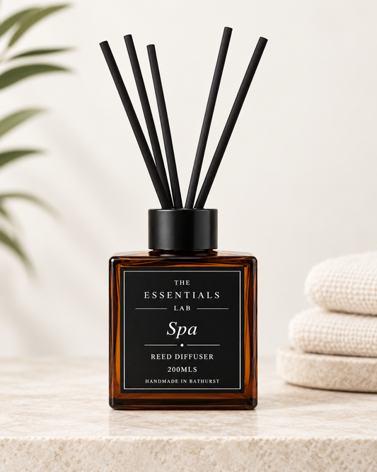 Amber glass reed diffuser bottle with a black cap and black reeds, labeled 'The Essentials Lab Spa Reed Diffuser 200mL', set on a light stone surface with a blurred plant and folded cream towels in the background.