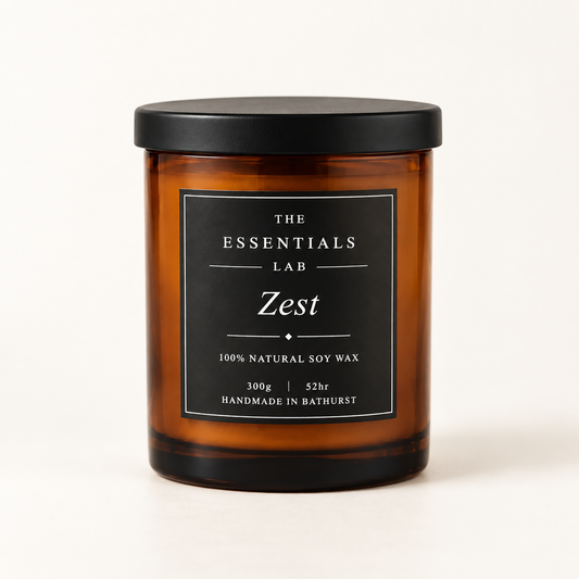 An amber glass jar candle with a black lid and a black label reading 'THE ESSENTIALS LAB Zest' on a clean white background.