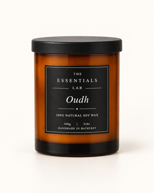 An amber glass jar candle with a black lid and a black label reading 'THE ESSENTIALS LAB Oudh' on a clean white background.
